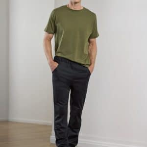 A man stands wearing olive green t-shirt and black jogger pants, with hands in pockets. The setting is minimalistic.