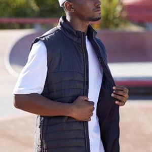 A man wears a black men's expedition vest, standing outdoors, with a white cap and t-shirt. The vest is unzipped.