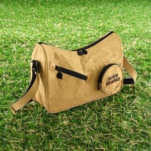 A tan Kraft paper sling bag with black accents and a round pocket featuring a logo. Set against green grass.